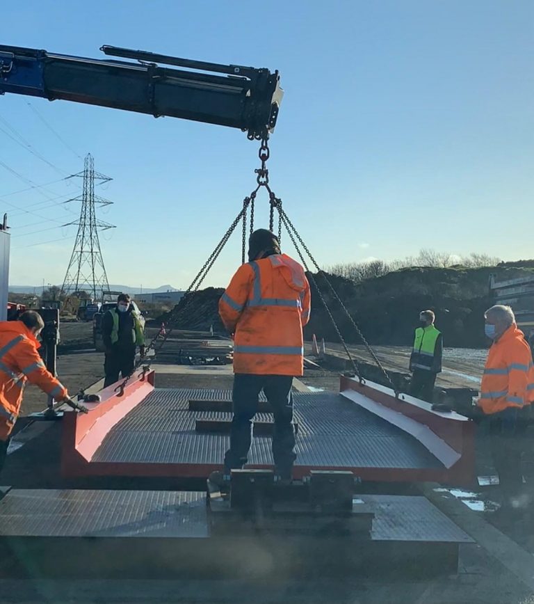 Four people in high visibility clothing wearing masks, standing at the corners of a large metal weighbridge being lowered from a crane.